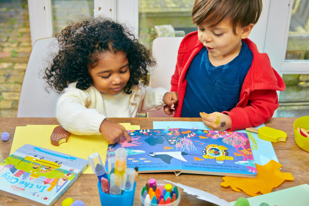 An image of two young children, a girl and a boy, sitting at table reading and interacting with a book together