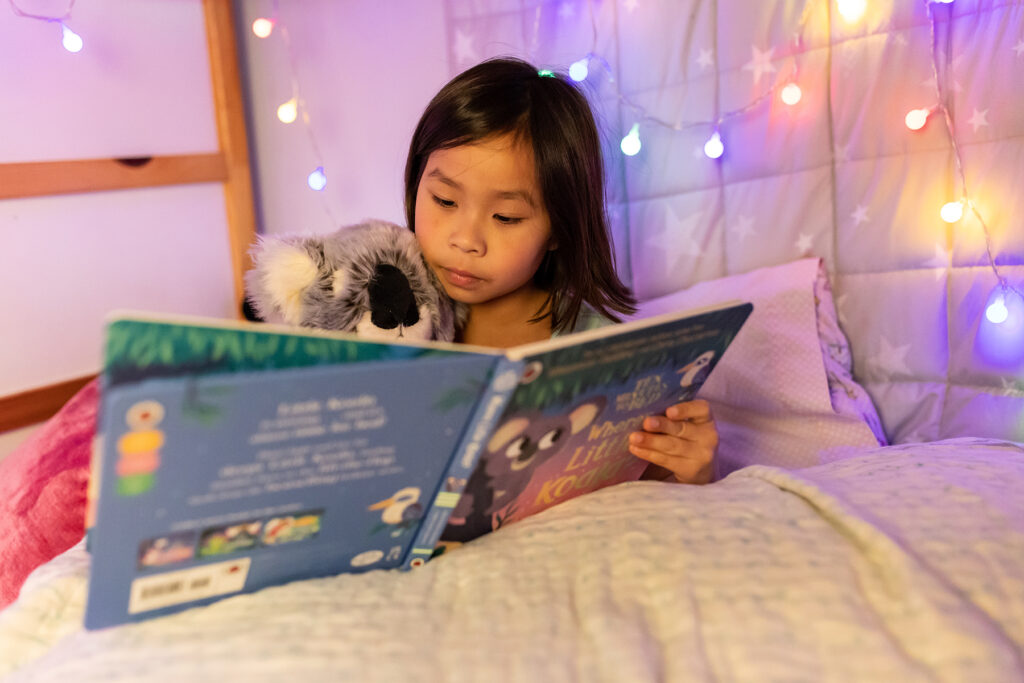 A photo of a little girl sitting in her bed reading. She is reading a story about a koala with her koala stuffed toy.