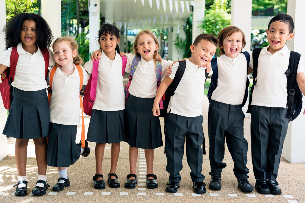 A photo of seven young children in their first year at school. They are standing and smiling for the camera with their arms around each other